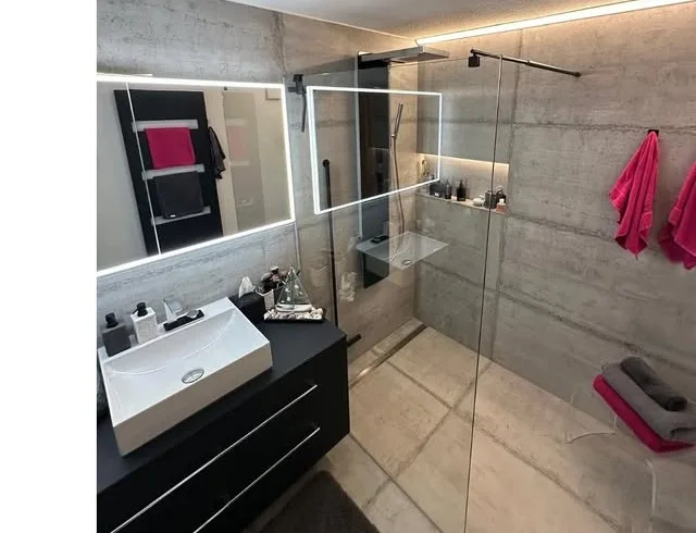 Fertiges Bad mit begehbarer Dusche hinter einer Glaswand, Waschtisch mit schwarzem Unterbau und Spiegel mit umlaufender Lichtkante
